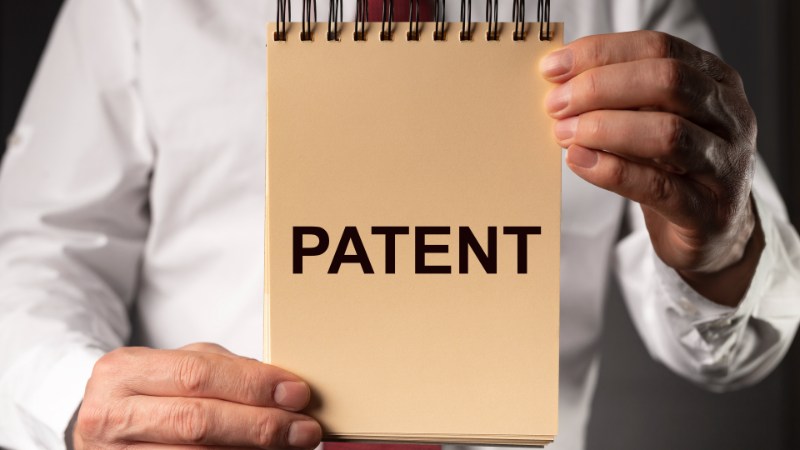 Uma pessoa de camisa branca e gravata vermelha segura um bloco de notas espiralado com as duas mãos. O bloco de notas tem uma capa bege com as palavras "PATENT - O Que São Ativos Intangíveis" impressas com destaque em letras maiúsculas pretas. O fundo escuro faz o bloco de notas se destacar claramente. - Efacont