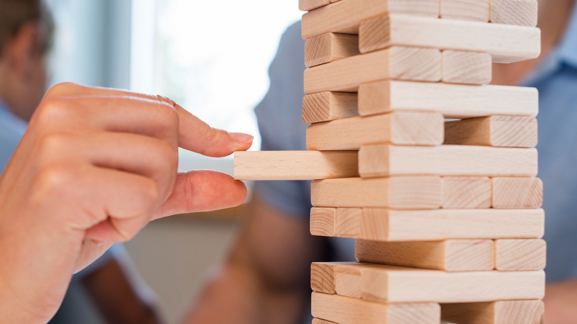 Um close de uma mão puxando cuidadosamente um bloco de madeira de uma torre Jenga. A torre consiste em blocos de madeira retangulares empilhados. O fundo está levemente desfocado, mostrando outros jogadores sutilmente fora de foco, indicando um ambiente interno — muito parecido com as complexidades de entender "O que é Balanço Patrimonial". - Efacont