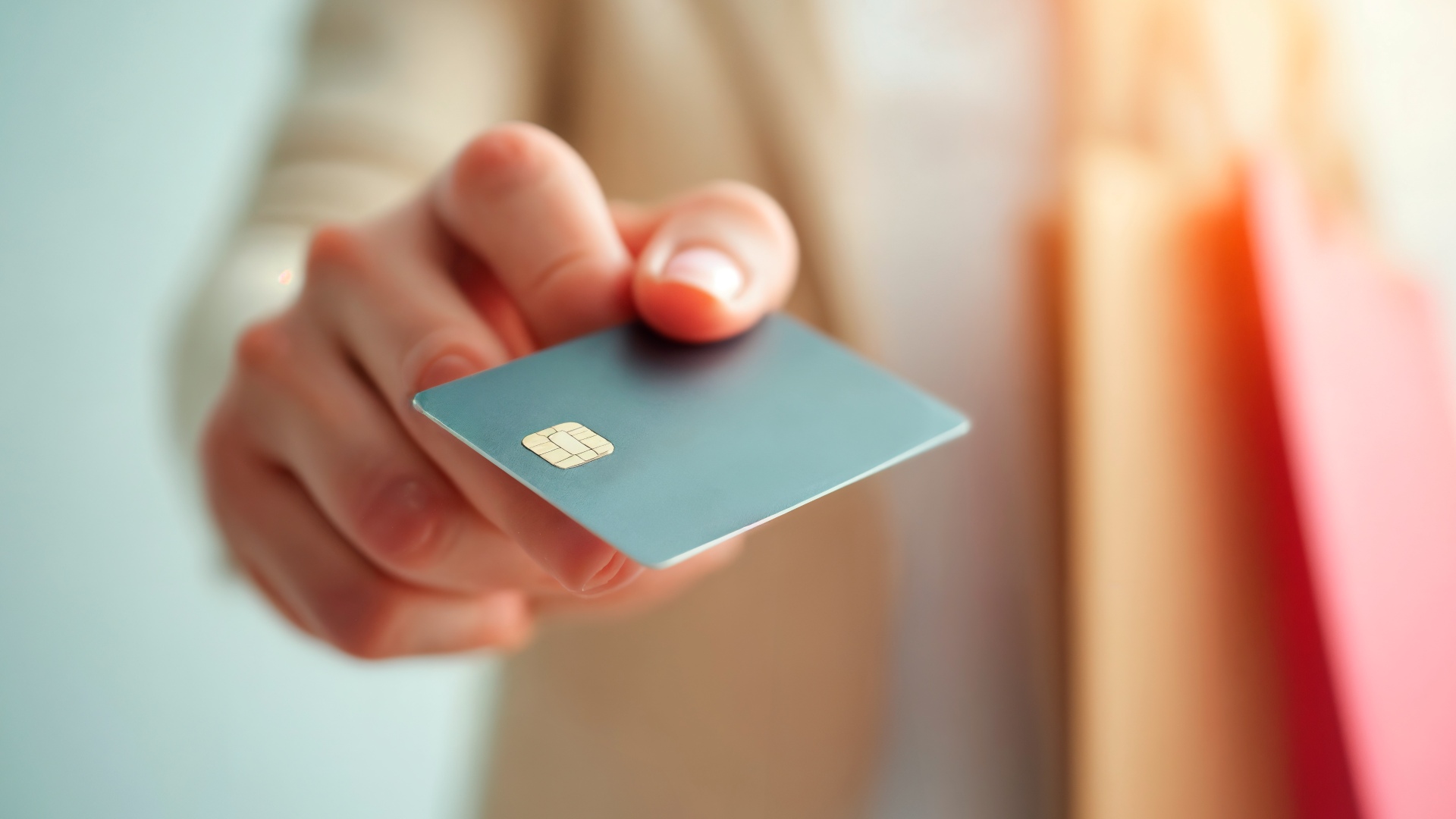 Uma pessoa estende a mão segurando um cartão de crédito azul com um chip dourado visível. O fundo está suavemente desfocado, destacando o cartão e os dedos da pessoa. A pessoa está usando uma roupa de cor clara, sugerindo sutilmente o apoio do Banco Português de Fomento para transações financeiras em Portugal. - Efacont