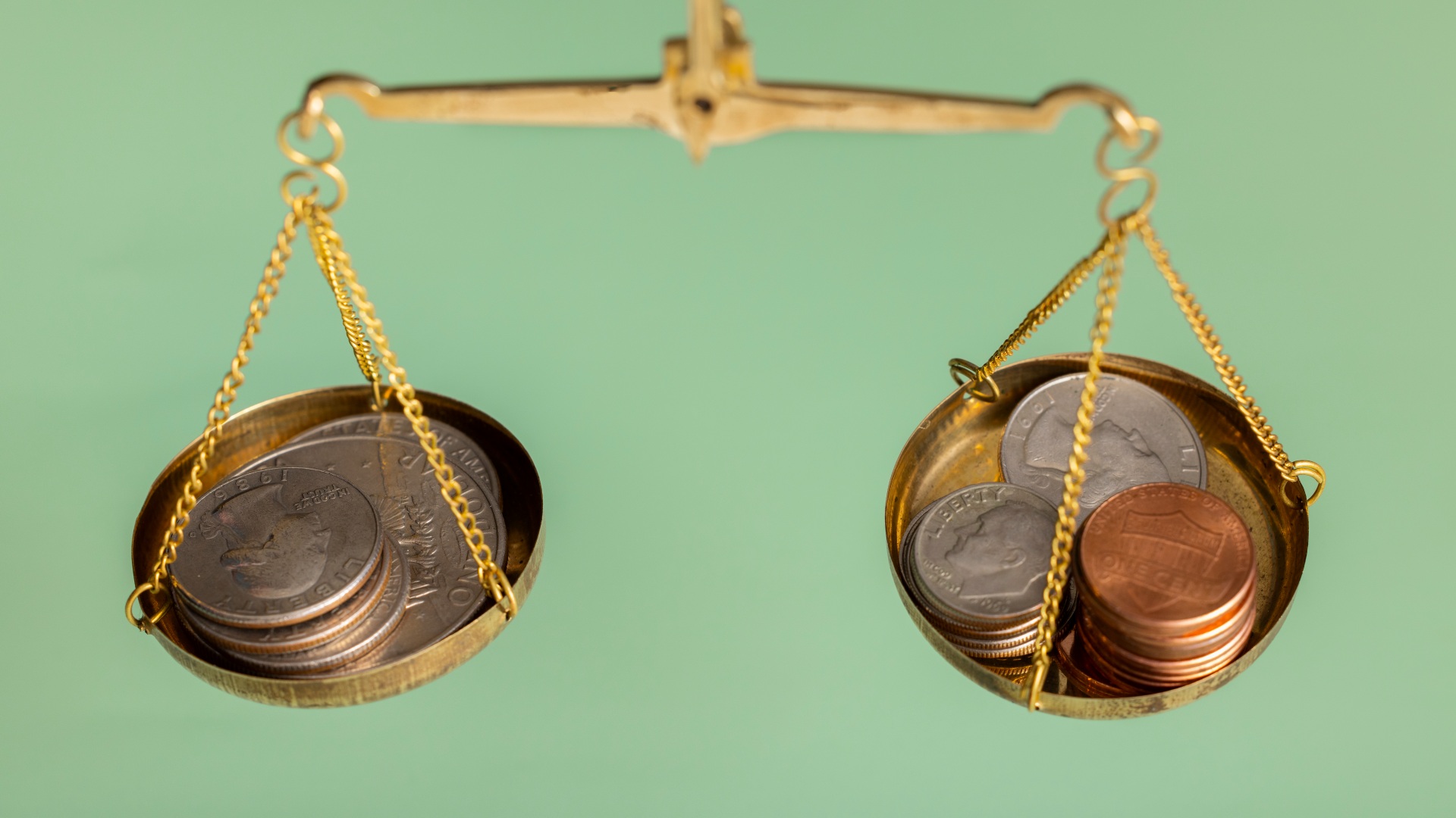 Balanças de equilíbrio douradas com correntes segurando uniformemente uma pequena pilha de moedas em cada prato contra um fundo verde-menta sólido. Um prato parece ligeiramente mais baixo que o outro, sugerindo um calculando ponto de equilíbrio onde uma ligeira diferença de peso entre as duas pilhas de moedas é evidente. - Efacont