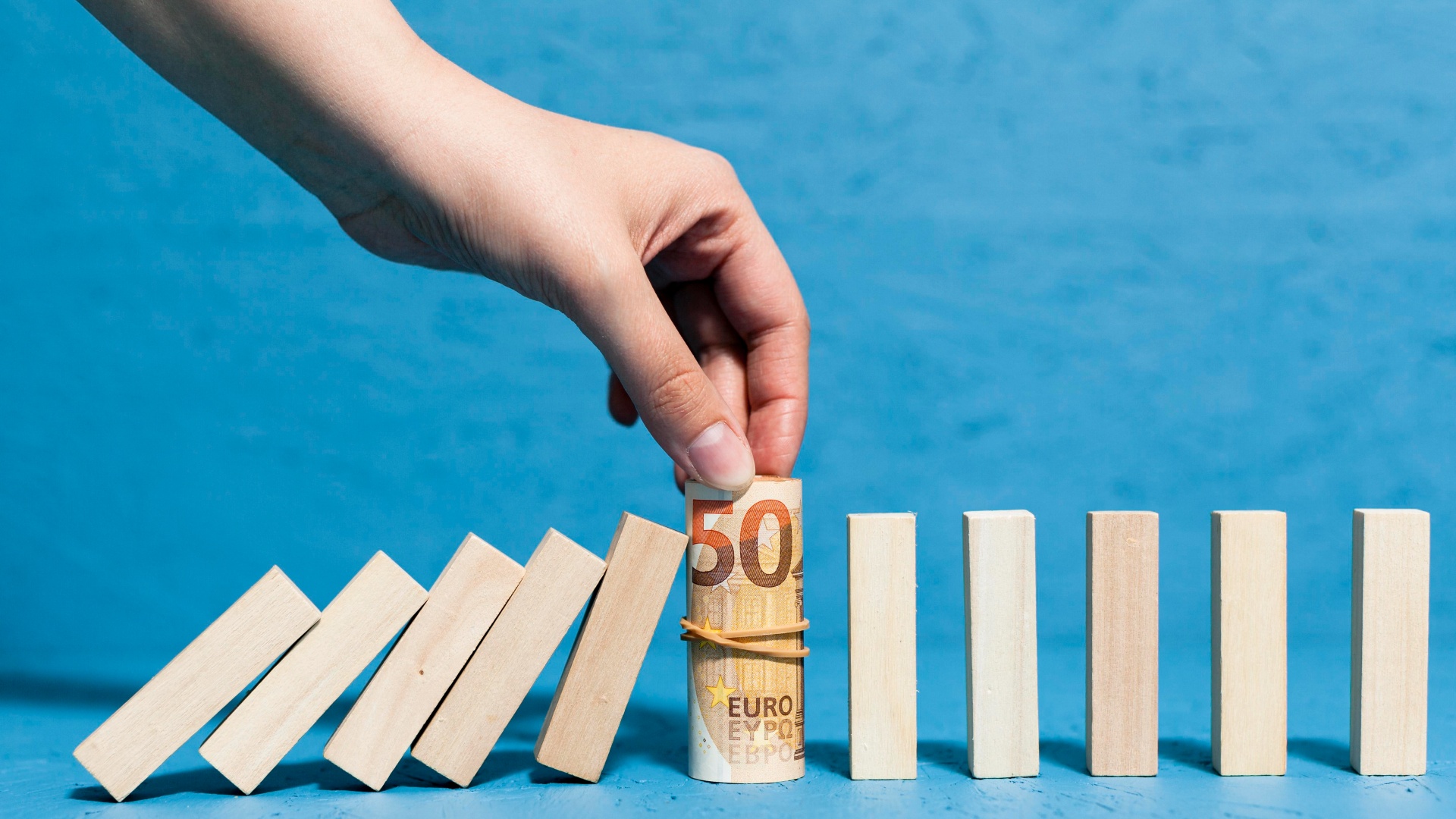 Uma mão é vista parando uma fileira de dominós de madeira caindo com uma nota de 50 euros enrolada, ecoando o conceito de "O que é Bailout", segurada entre o polegar e os dedos. A nota de euro é presa com um elástico contra um fundo azul, criando um contraste marcante com os dominós e a moeda de cor clara. - Efacont