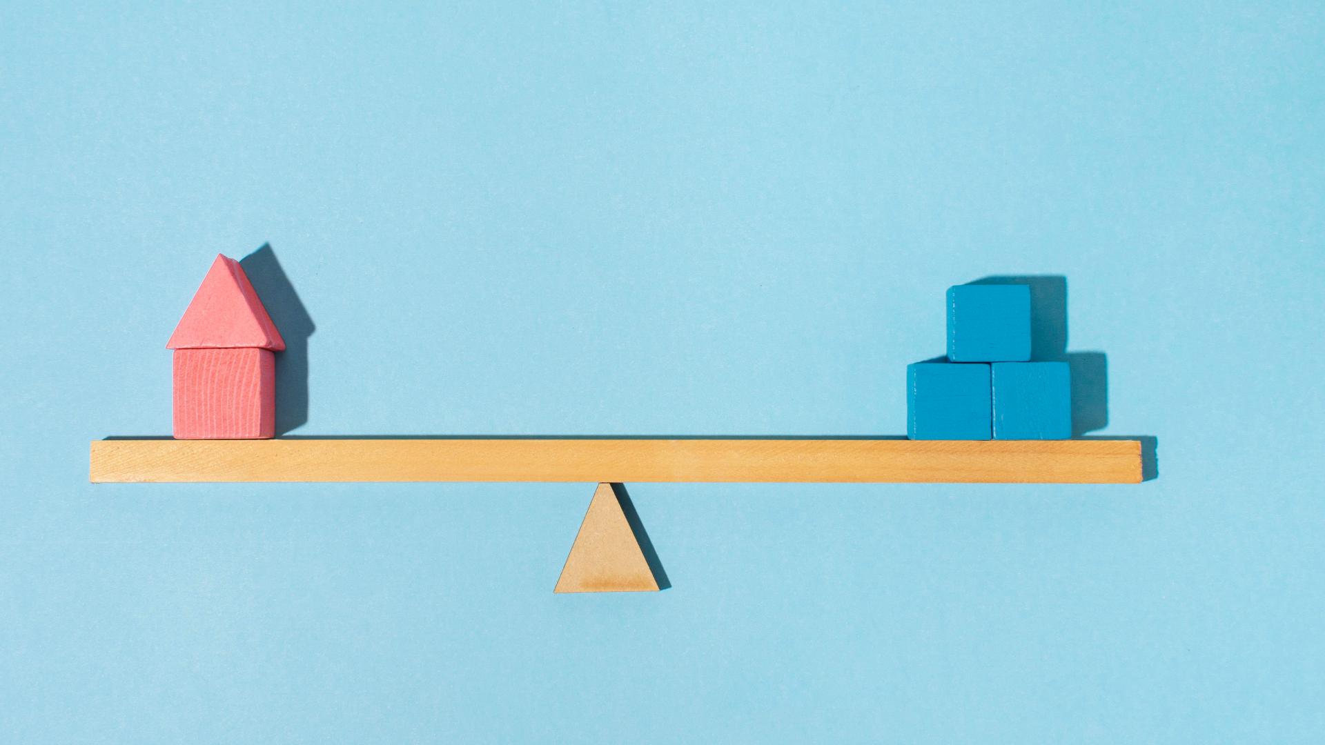 Uma gangorra de madeira equilibrada contra um fundo azul claro. No lado esquerdo da gangorra há um bloco de madeira rosa no formato de uma casa, e no lado direito há três pequenos cubos de madeira azuis empilhados em dois níveis. A gangorra e os objetos criam um equilíbrio visual. - Efacont