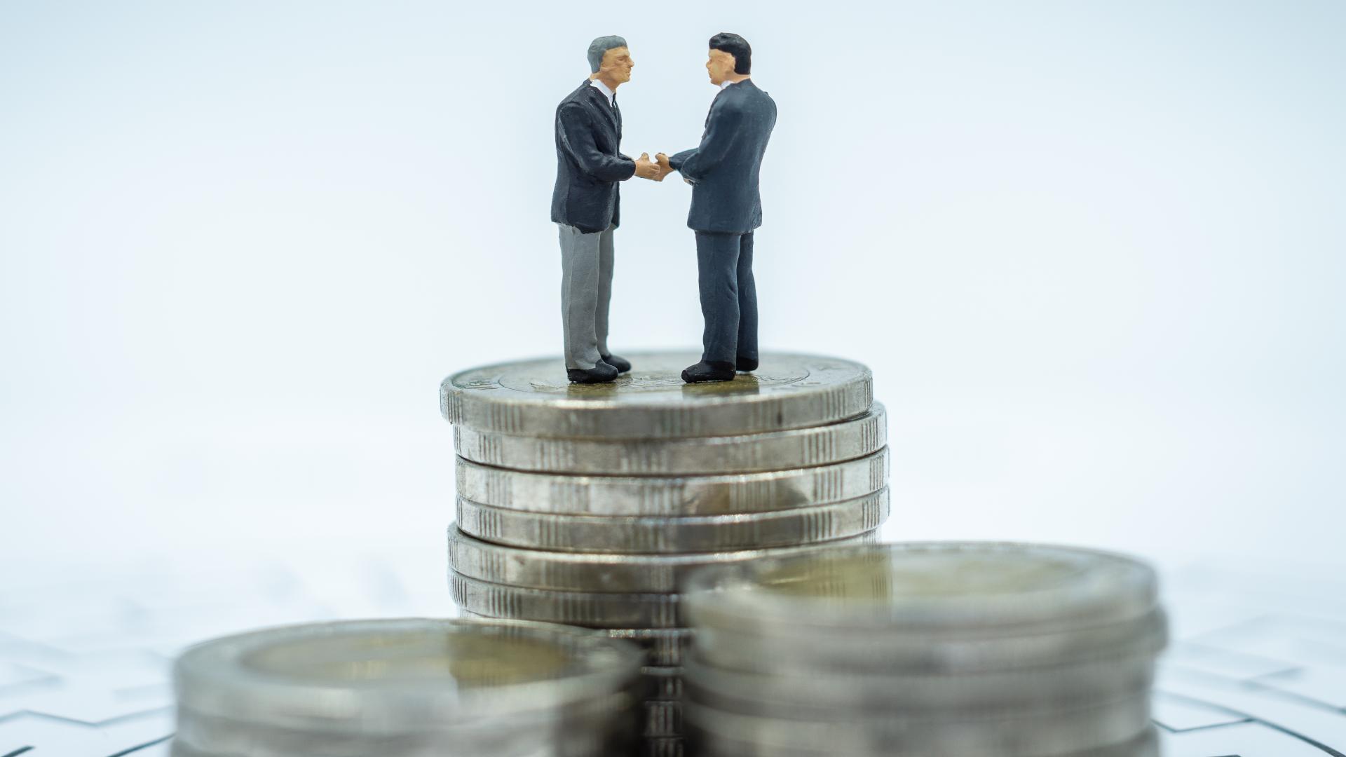 Duas figuras em miniatura de homens de terno apertando as mãos ficam em cima de uma pilha de moedas, simbolizando o significado de um acordo ou negócio. O fundo suavemente desfocado com pilhas de moedas adicionais enfatiza o tema de transações financeiras e capital subscrito. - Efacont