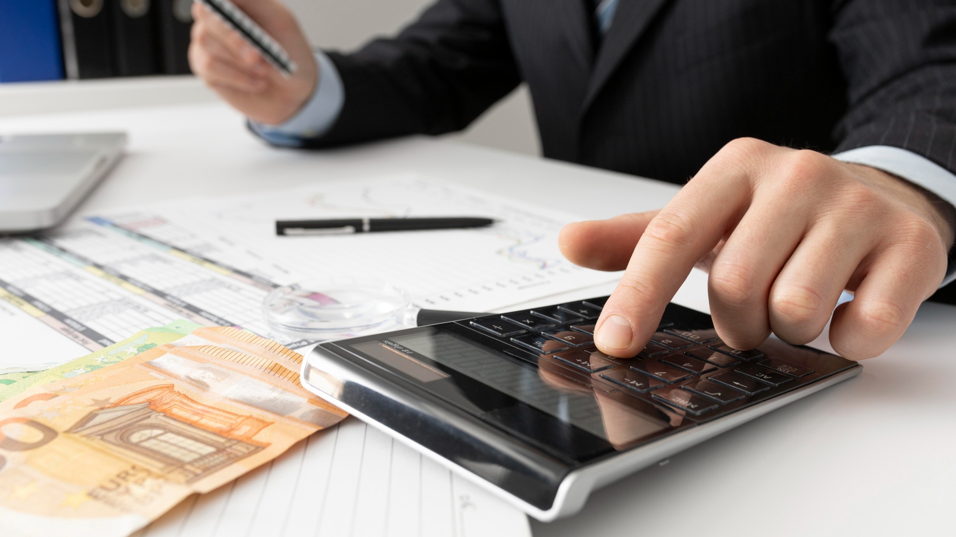 Uma pessoa de terno usa uma calculadora em uma mesa abarrotada de documentos, uma lupa, uma caneta e várias notas de Euro. A mão da pessoa está pressionando um botão na calculadora enquanto segura um telefone, possivelmente pesquisando o que é capital alheio como parte de suas atividades de análise financeira ou orçamento. - Efacont