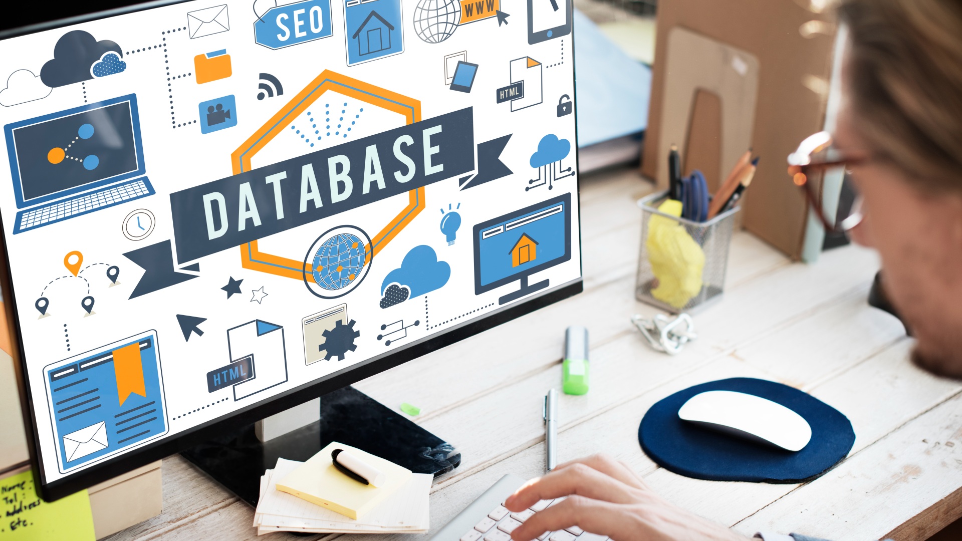 Uma pessoa trabalhando em uma mesa com um computador exibindo um gráfico sobre marketing de banco de dados. A tela mostra vários ícones relacionados à tecnologia, como uma nuvem, cadeado e engrenagens, junto com a palavra "DATABASE" em letras grandes. A mesa contém um teclado, mouse, notebook e itens de papelaria. - Efacont