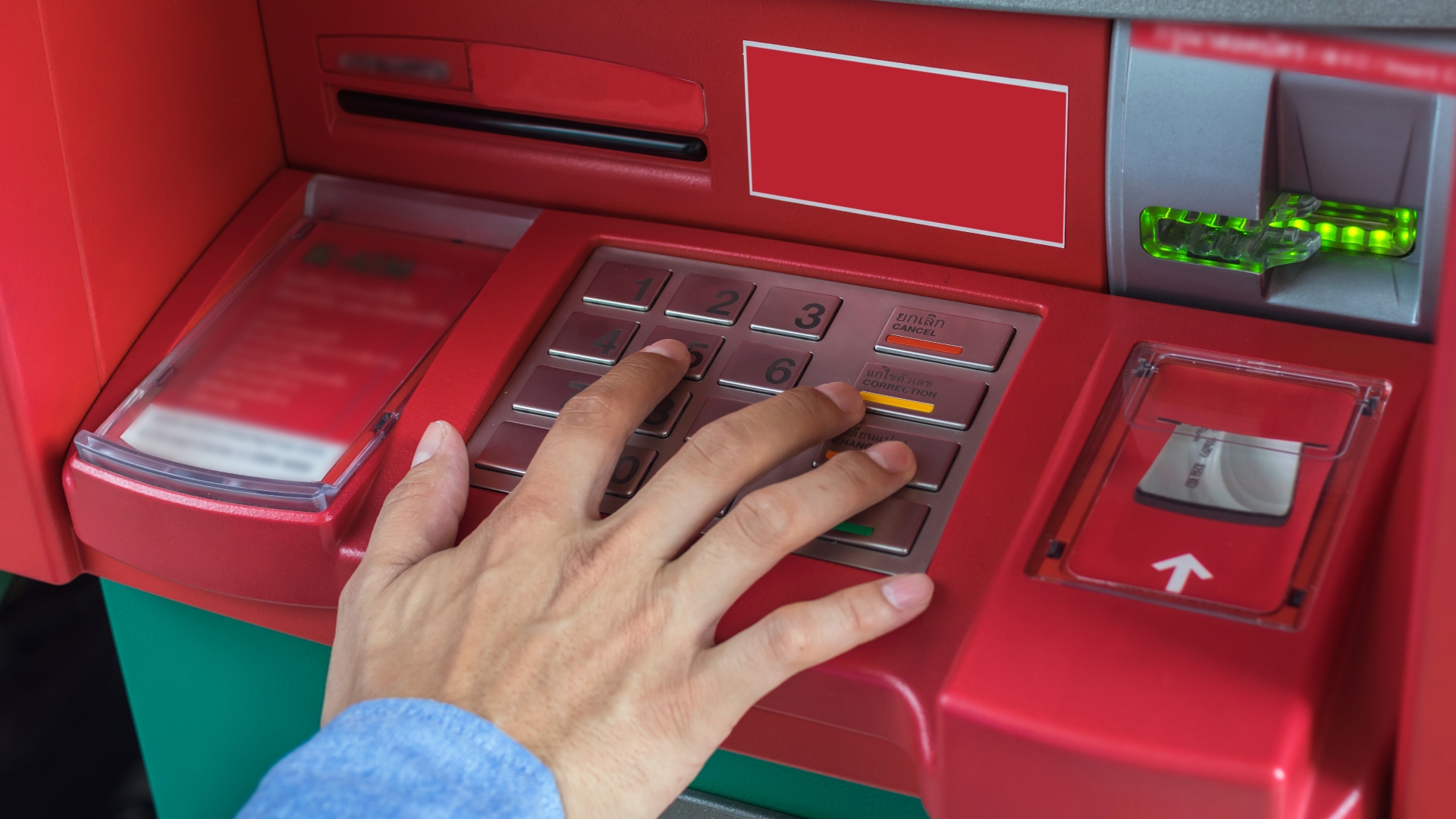 Uma imagem em close-up mostra a mão de uma pessoa, com uma manga azul, pressionando botões em um teclado de caixa eletrônico. O caixa eletrônico vermelho com detalhes em verde e cinza apresenta um slot para cartão e uma tela exibindo instruções em inglês e espanhol, perfeito para usuários curiosos sobre descoberto bancário o que é. - Efacont
