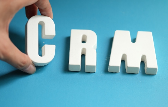 Uma mão segura uma letra maiúscula branca "C" ao lado das letras maiúsculas "R" e "M", todas em uma superfície azul, formando "CRM", representando Software de Gestão para Relacionamento com o Cliente. - Efacont