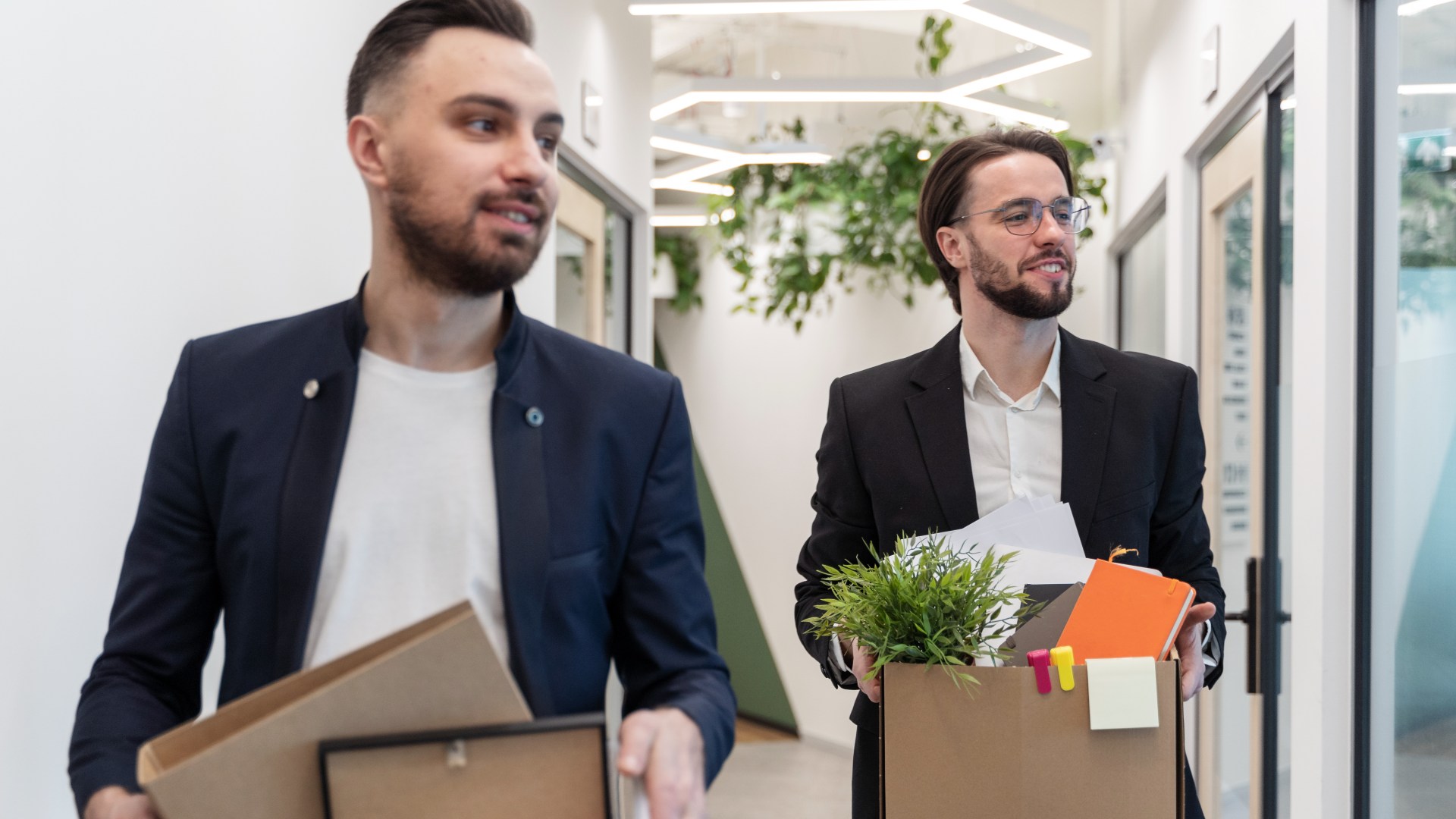 Dois homens em trajes de negócios caminham por um corredor de escritório, cada um carregando uma caixa cheia de itens pessoais, como uma planta em vaso, livros e artigos de papelaria. O espaço bem iluminado reflete os recentes esforços de redução de pessoal, com luzes de teto modernas e portas de vidro de escritório alinhando seu caminho. - Efacont