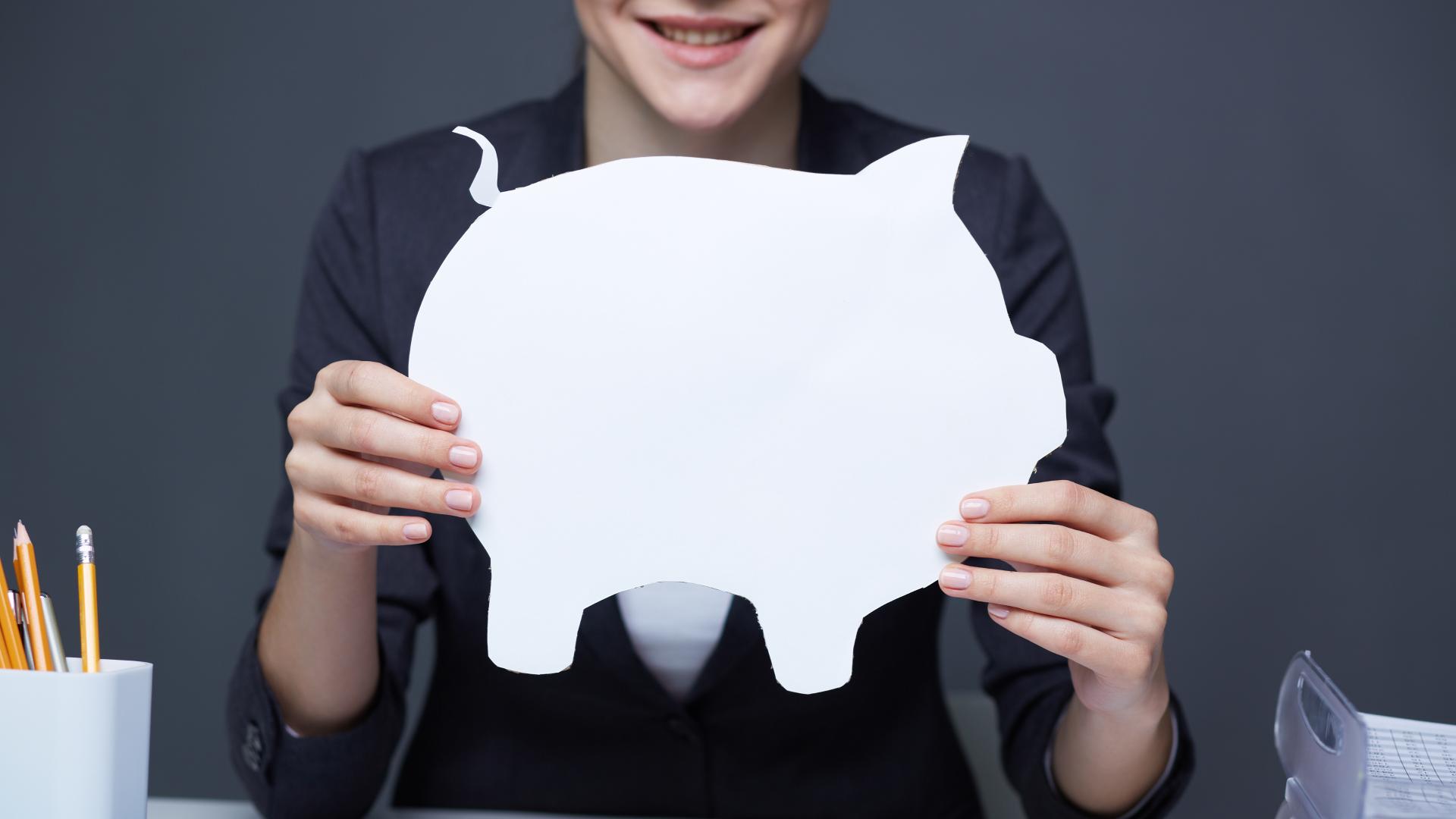 Uma pessoa segurando um grande recorte de papel branco de um cofrinho na frente do peito. A pessoa está sorrindo, vestindo um blazer escuro, incorporando o espírito do investimento. O fundo é cinza, e uma xícara com lápis e uma pilha de papéis são visíveis na mesa, sugerindo seu grande interesse em finanças. - Efacont