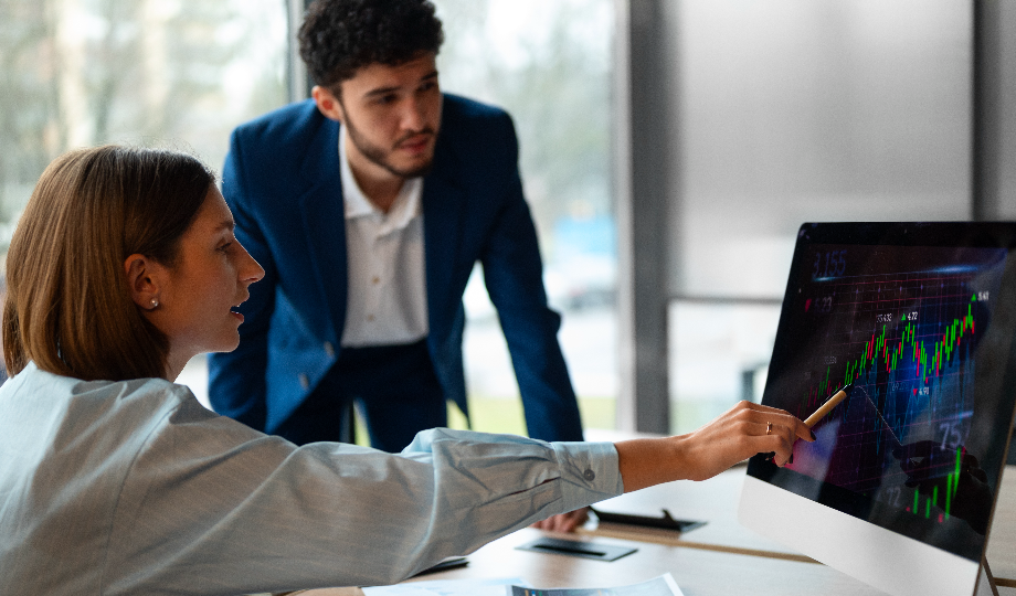 Uma mulher aponta para uma tela de computador exibindo um gráfico colorido do Mercado Financeiro, enquanto um homem de terno azul se inclina para observar. Ambos parecem concentrados e engajados, sentados em um escritório moderno e iluminado, com grandes janelas atrás deles. - Efacont