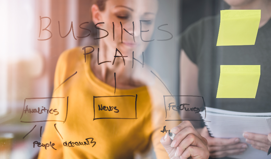 Uma mulher escreve "BUSSINES PLAN" em um quadro de vidro com seções de fluxograma rotuladas como "Analytics", "News" e "Features" - ilustrando o que é plano de negócios. Ela segura um marcador, enquanto outra pessoa segura papéis. Duas notas adesivas amarelas estão sobre o vidro. - Efacont