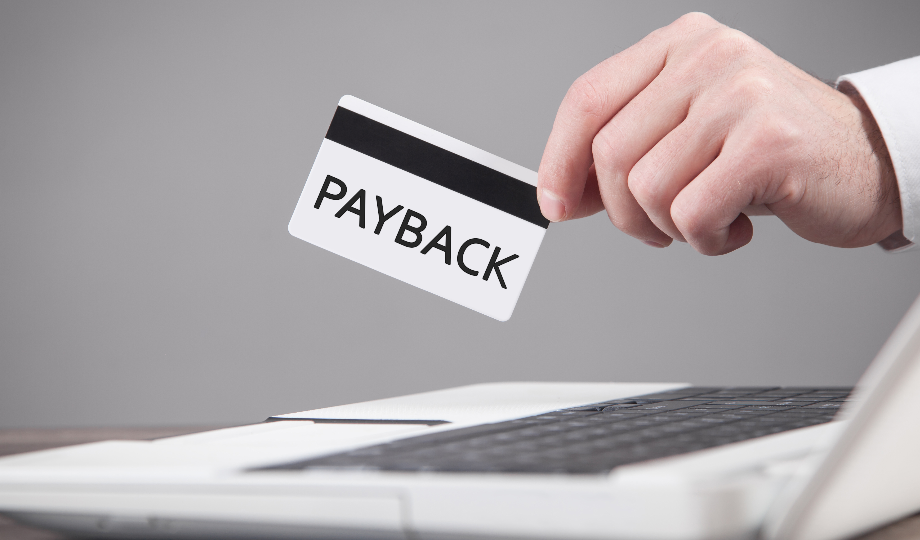 Uma mão segura um cartão branco com a inscrição "PAYBACK" acima de um teclado de laptop, destacando o conceito de cálculo de payback ou reembolso financeiro por meio de métodos digitais. O fundo liso e desfocado enfatiza o cartão e o laptop. - Efacont