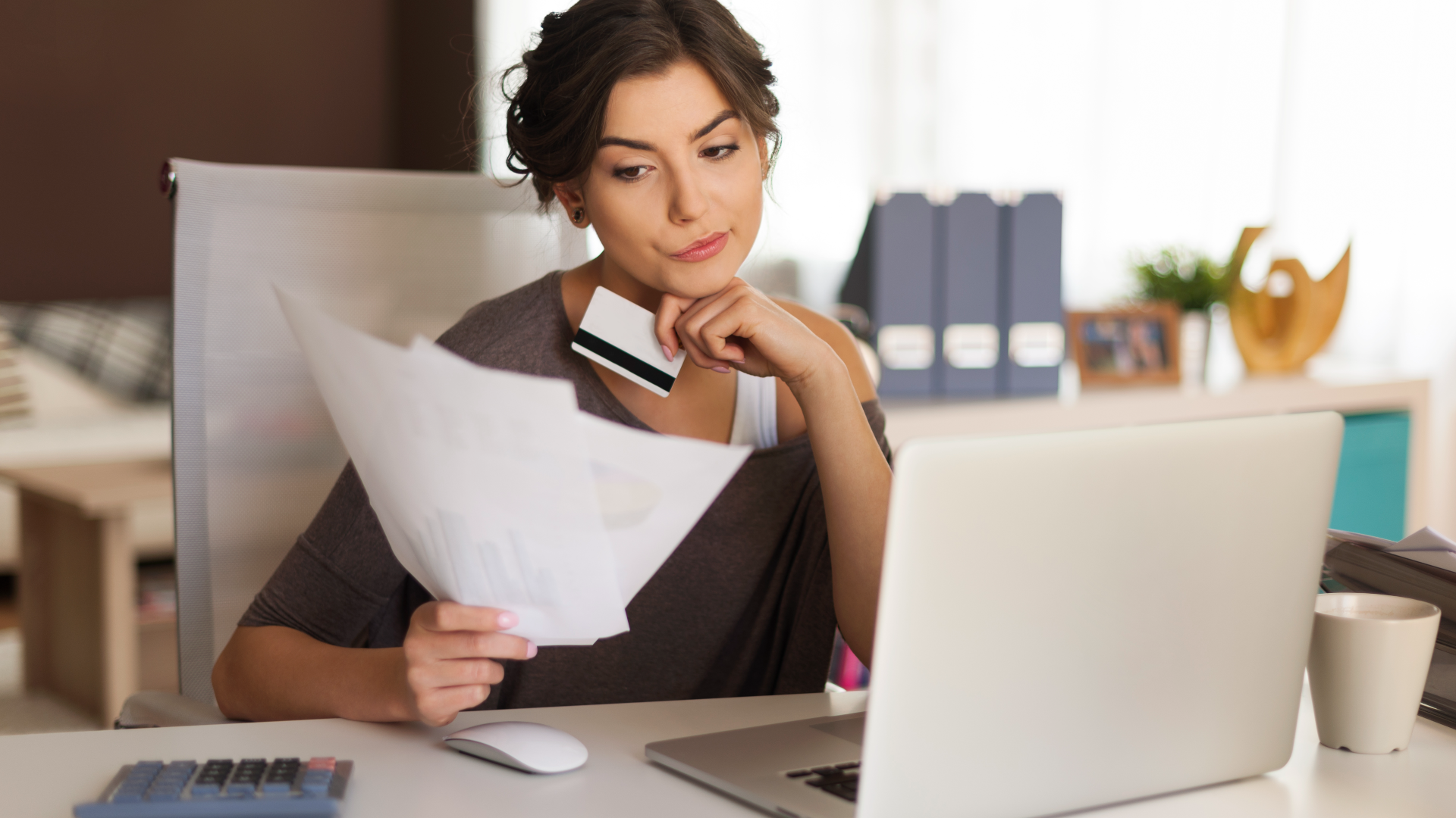 Uma mulher está sentada em uma mesa, segurando um cartão de crédito e papéis enquanto olha pensativamente para uma tela de laptop exibindo um artigo sobre "O que São Empresas Zumbi". Ela veste um suéter marrom, e uma xícara e uma calculadora estão sobre a mesa. Uma luz natural suave preenche a sala, destacando uma prateleira com fichários e itens decorativos atrás dela. - Efacont