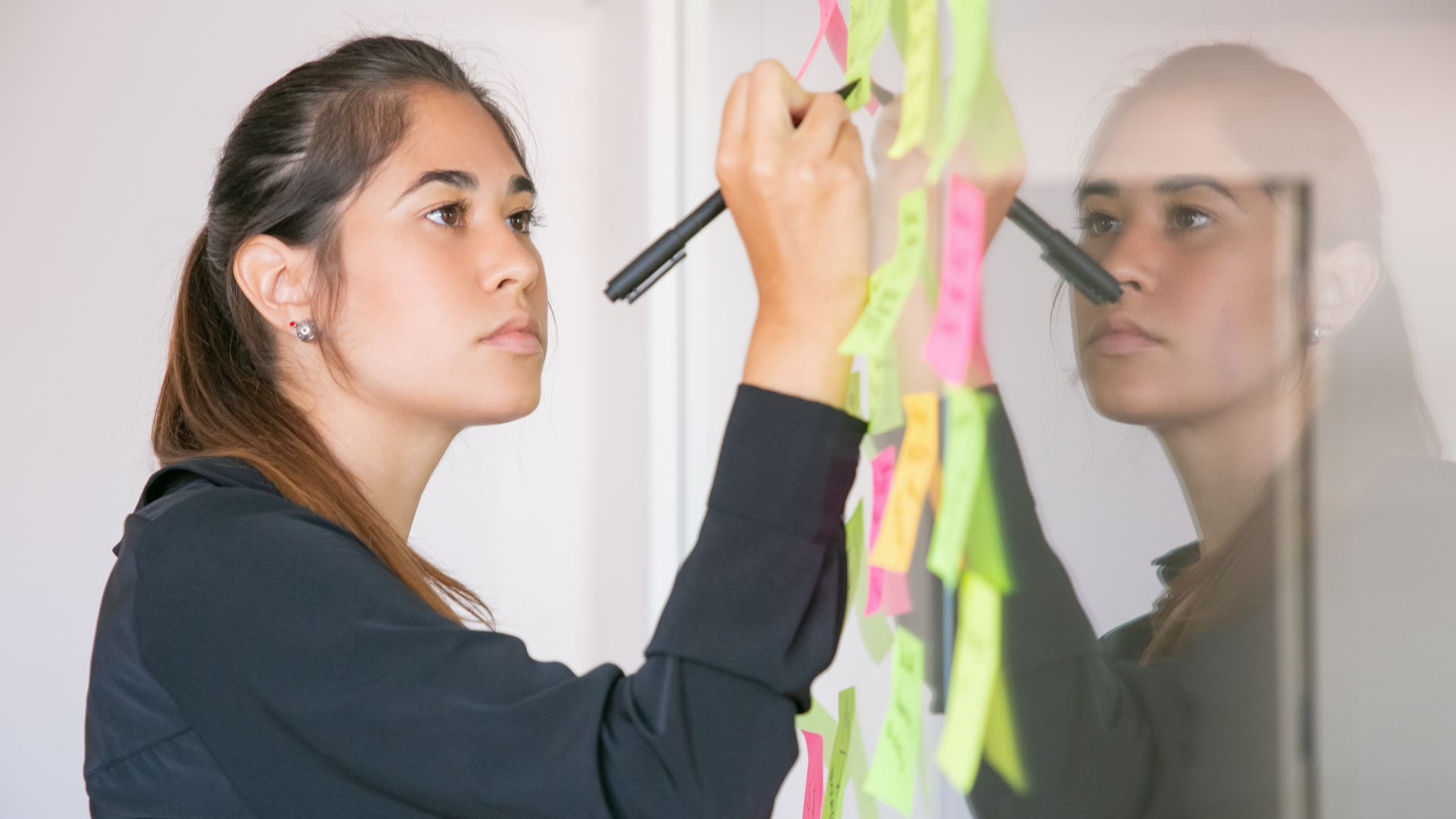 Uma mulher com longos cabelos castanhos está imersa na escrita "O Que é Fail Fast" em post-its coloridos presos a uma parede de vidro. Usando um marcador preto, ela cria um padrão organizado com notas rosa, amarelas e verdes. Seu reflexo brilha no vidro enquanto seu top preto e brincos completam a cena. - Efacont