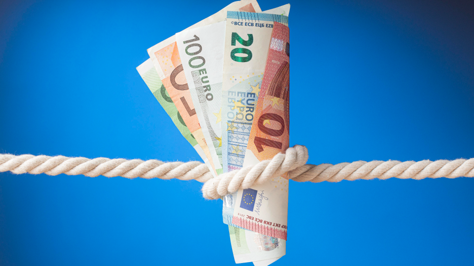 Um maço de diversas notas de euro, incluindo notas de 10, 20 e 100 euros, está firmemente preso por uma corda grossa sobre um fundo azul sólido, simbolizando inflação econômica e pressão ou restrição financeira. - Efacont