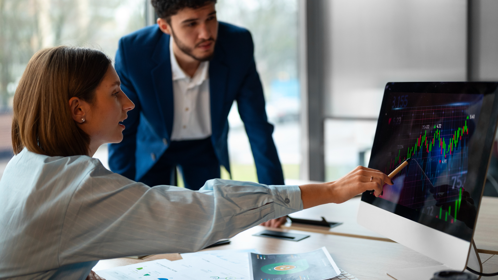 Uma mulher e um homem trabalham juntos em um ambiente de escritório, analisando um gráfico financeiro em um monitor de computador. A mulher aponta para a tela com uma expressão focada, discutindo mutualmente exclusivos investimentos. Papéis estão espalhados na mesa, enquanto o homem se inclina, observando atentamente. - Efacont