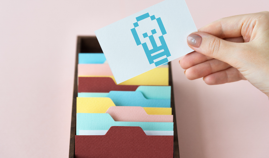 Uma mão segura um cartão branco com uma lâmpada azul pixelizada acima de uma caixa aberta de fichas coloridas — perfeitas para definição de metas ou organização de objetivos SMART — contra um fundo rosa suave. - Efacont