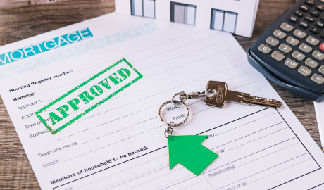 Close-up de um formulário de solicitação de hipoteca carimbado com "APROVADO" em tinta verde. Uma chave de casa em um chaveiro verde em forma de seta repousa sobre o formulário, sugerindo um financiamento de arrendamento bem-sucedido para a compra de uma casa. Uma calculadora e parte de um documento são visíveis ao fundo. - Efacont