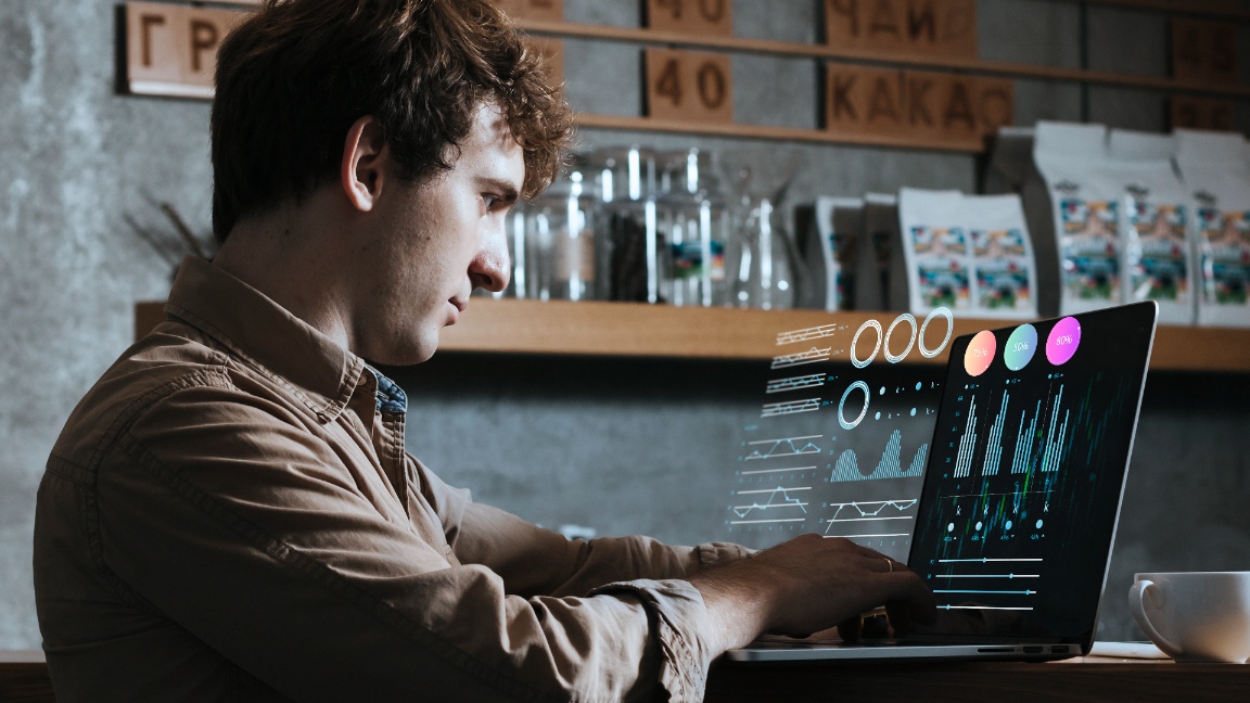 Um homem de camisa bege trabalha em um laptop em um café moderno, a tela exibindo gráficos e tabelas digitais futuristas — um aceno para estratégias de growth hacking. Prateleiras cheias de equipamentos e bolsas de café criam um ambiente aconchegante e industrial. - Efacont