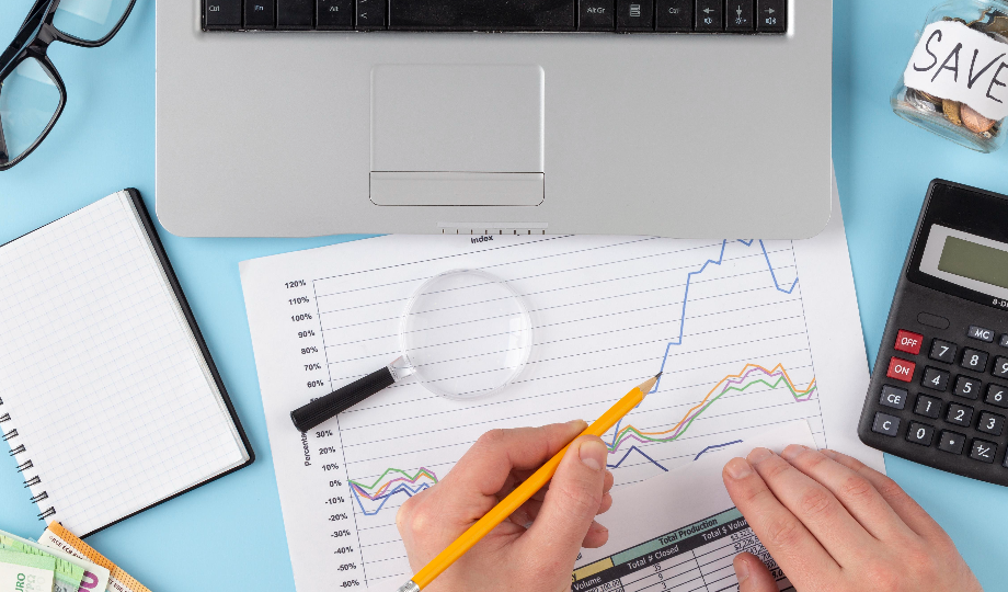 Uma pessoa analisa um gráfico financeiro com um lápis ao lado de um laptop, uma calculadora, uma lupa, um caderno e um pote com o rótulo "SAVE" em uma mesa azul - ilustrando o planejamento financeiro e os fundamentos das finanças pessoais. - Efacont