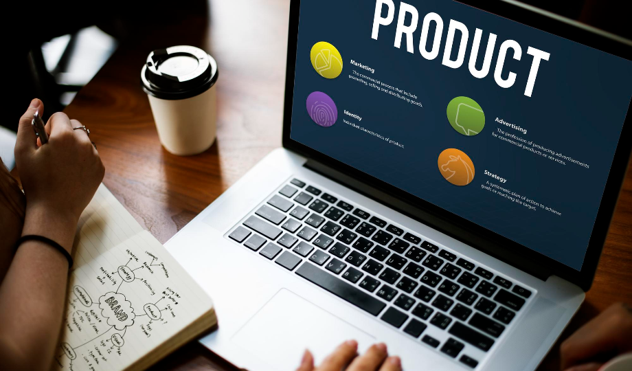 Uma pessoa trabalhando em um laptop mostra um slide intitulado "PRODUTO" com quatro ícones coloridos: Produção, Marketing, Publicidade e Estratégia. Ao lado deles, há um caderno cheio de notas manuscritas e um mapa mental de matriz BCG, junto com uma xícara de café para viagem na mesa de madeira. - Efacont