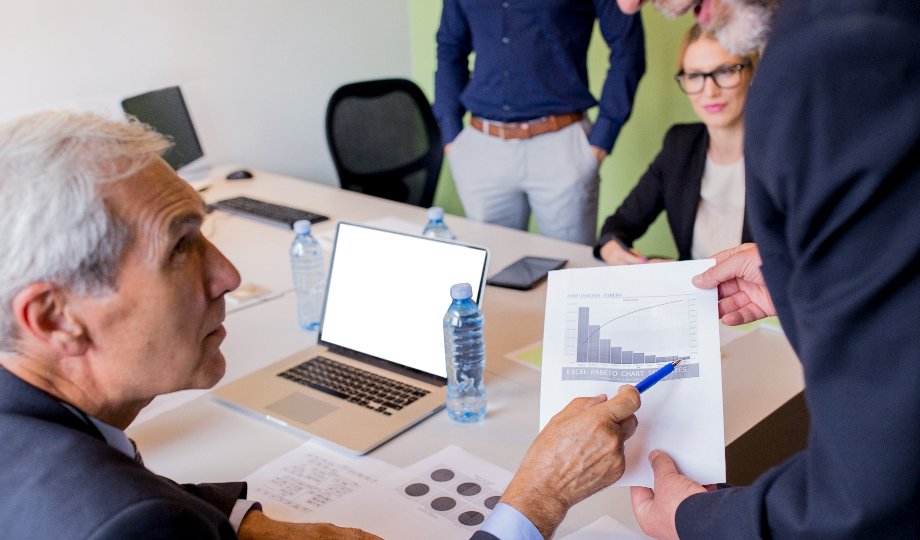 Quatro profissionais se reúnem em uma mesa com laptops e papéis. Um deles aponta para um gráfico, discutindo preços de transferência, enquanto outro ouve atentamente. A atmosfera concentrada sugere uma discussão comercial detalhada ou uma apresentação sobre preços de transferência. - Efacont