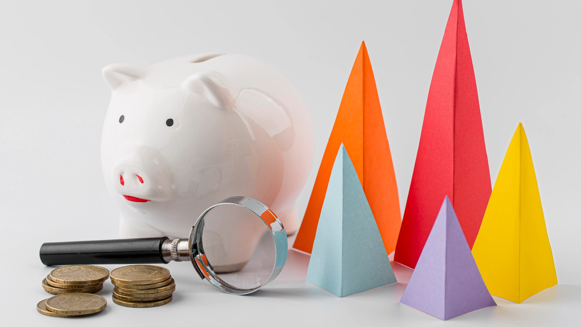 Um cofrinho branco ao lado de uma pilha de moedas e uma lupa fica à esquerda, enquanto à direita, quatro vibrantes pirâmides de papel simbolizam crescimento financeiro. Este arranjo convida à curiosidade sobre "o que é Grau de abertura na economia" contra um fundo simples e claro. - Efacont