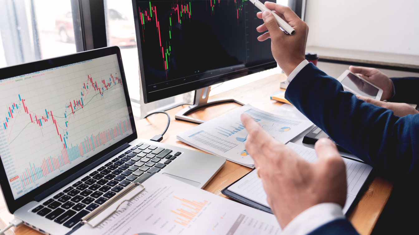 Duas pessoas de terno analisam gráficos financeiros em um laptop e dois monitores, discutindo estratégias como "O que é Hedge". A mesa contém papéis com gráficos e um smartphone. Uma pessoa aponta para uma tela com uma caneta, enquanto a luz do dia entra por uma janela, destacando seu ambiente colaborativo. - Efacont