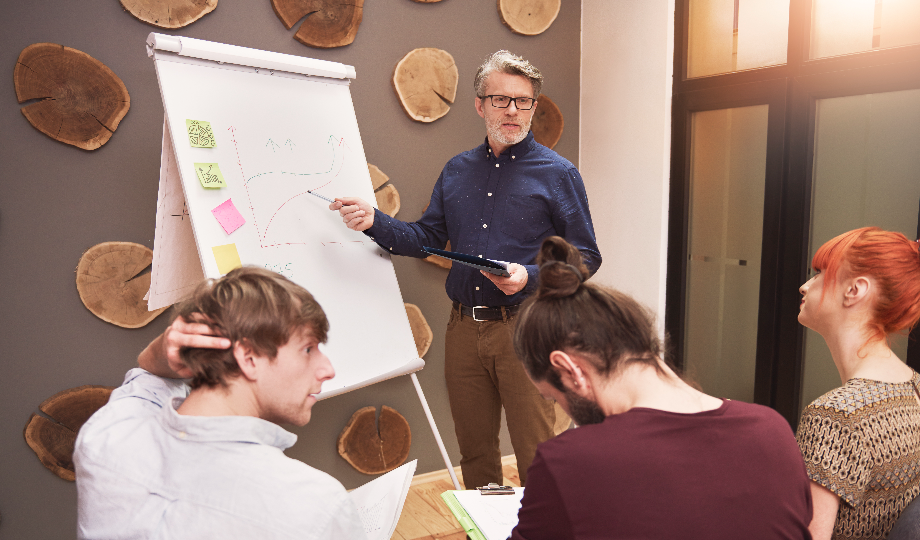 Um homem está em pé na frente de um flip chart, apontando para um gráfico durante sua apresentação sobre a diferença entre coaching e mentoring. Três pessoas sentam-se de frente para ele: um homem tocando seu cabelo, outro com um coque tomando notas e uma mulher com cabelo vermelho. A sala ostenta detalhes em madeira e uma grande janela. - Efacont