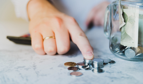 Uma mão usando um anel está empilhando moedas sobre uma mesa, simbolizando o conceito de "O que é Liquidez Geral". Ao lado, um jarro de vidro transparente contém mais moedas e algumas notas de papel dobradas. O fundo suavemente desfocado enfatiza a atividade financeira em primeiro plano. - Efacont