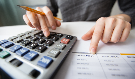 Uma pessoa, vestindo uma camisa cinza, usa uma calculadora grande com um lápis na mão enquanto a outra mão aponta para números em uma planilha. A imagem sugere trabalho financeiro, possivelmente envolvendo estratégias para lidar com flutuações no mercado financeiro. - Efacont
