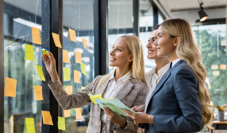 Três mulheres em trajes de negócios colaboram em um escritório, colocando notas adesivas coloridas em uma parede de vidro, que lembra um quadro kanban. Elas estão envolvidas e sorrindo, segurando papéis e discutindo ideias. A luz natural filtra através de grandes janelas, criando um ambiente de trabalho moderno e brilhante. - Efacont