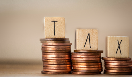 Blocos de madeira com as letras "T", "A" e "X" repousam sobre três pilhas de moedas de cobre, formando a palavra "TAX". As pilhas variam em altura contra um fundo bege, simbolizando conceitos financeiros como tributação ou ilustrando aspectos como juros de mora (juros sobre pagamentos em atraso). - Efacont