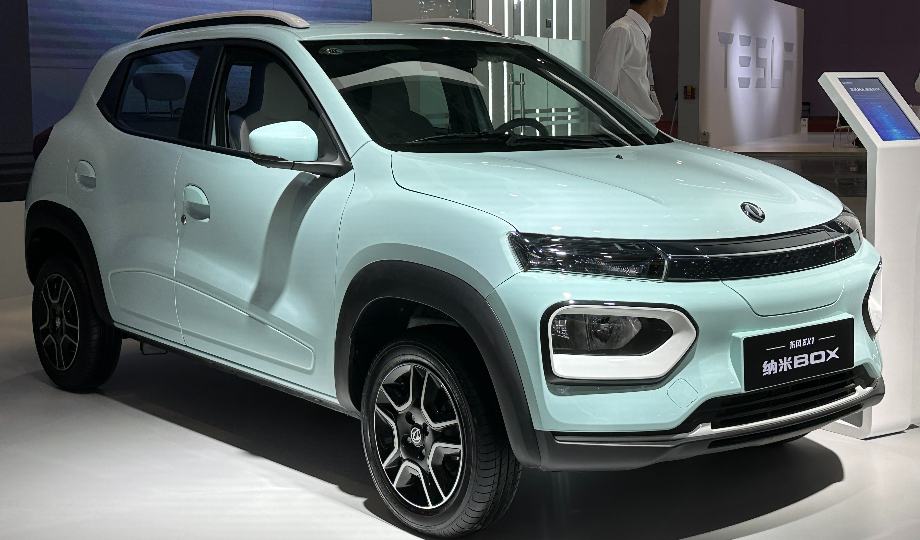 Um SUV compacto azul claro é exibido em ambientes internos. Com faróis elegantes e rodas de liga leve prateadas, este moderno veículo elétrico de 2025 ostenta excelente custo-benefício. Posicionado em uma plataforma branca ao lado de um estande interativo, o cenário inclui uma imagem desfocada e elementos de design abstratos. - Efacont