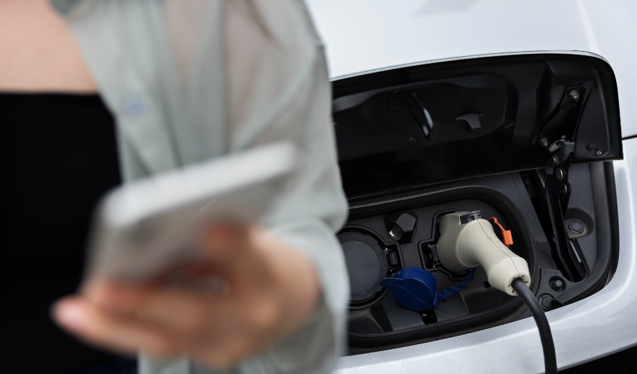 Uma pessoa segurando um smartphone está perto de um carro elétrico branco, com novas baterias elétricas, com uma porta de carregamento aberta. O cabo de carregamento está conectado. A pessoa veste uma camisa verde-clara sobre uma blusa preta, seu rosto desfocado e fora de foco. - Efacont