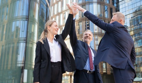 Três empresários em trajes formais estão dando high-fives ao ar livre em frente a modernos edifícios de vidro, personificando o espírito de 'empresas valiosas'. A mulher à esquerda usa um terno preto, enquanto os dois homens usam ternos escuros. Eles estão comemorando como se estivessem marcando uma história de sucesso per capita. - Efacont