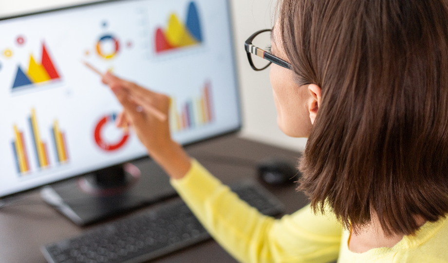 Uma mulher com óculos e cabelos castanhos está sentada em uma mesa, apontando com um lápis para um monitor de computador exibindo gráficos e tabelas coloridas. Ela veste um suéter amarelo, ilustrando "estudos de mercado o que é" por meio de gráficos de barras, linhas e pizza. Um teclado é visível na frente do monitor. - Efacont