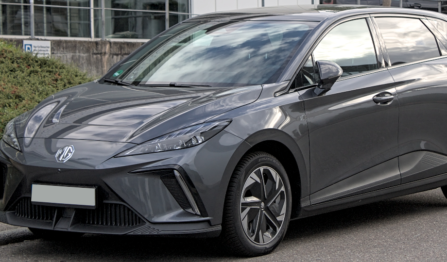 O carro elétrico MG cinza, um modelo 2025, está estacionado diagonalmente na rua, exibindo curvas elegantes e faróis angulares. Com seu design aerodinâmico moderno e grade frontal sutilmente integrada, ele incorpora um visual futurista — realmente um destaque no mundo dos carros elétricos. - Efacont