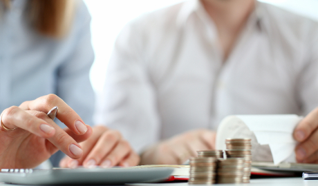 Duas pessoas sentam-se em uma mesa carregada com pilhas de moedas e uma calculadora, profundamente envolvidas em planejamento financeiro. Uma aperta botões da calculadora com uma caneta, enquanto a outra examina um longo recibo, ambas em camisas de cor clara, aparentemente discutindo as nuances do Regime de Iva de Caixa. - Efacont