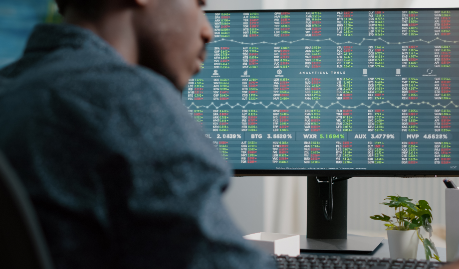 Uma pessoa de jaqueta azul observa uma grande tela de computador exibindo um painel colorido do mercado de ações, contemplando "O que é a Bolsa de Valores". Uma pequena planta e um caderno estão ao lado do monitor no moderno e bem iluminado escritório. - Efacont