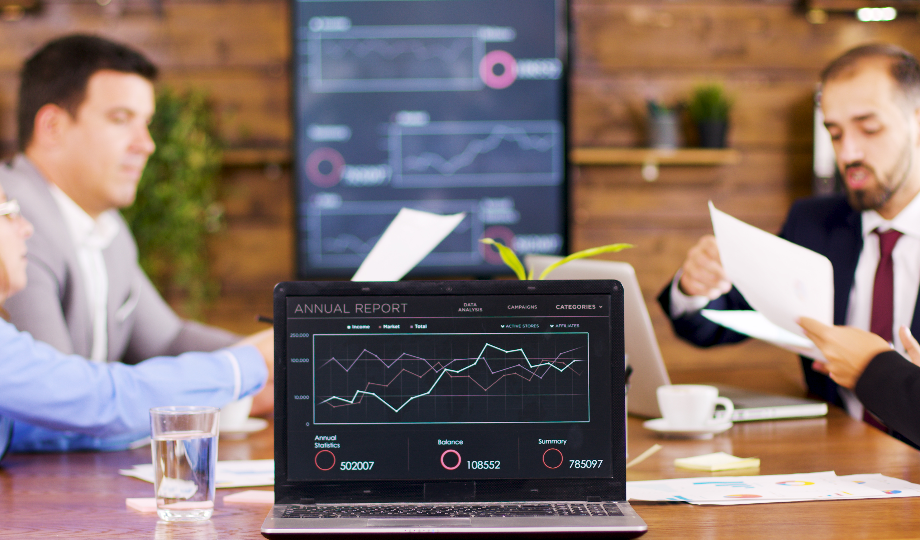 Quatro pessoas em trajes executivos sentam-se ao redor de uma mesa com documentos e laptops. Um laptop em primeiro plano exibe um relatório anual intitulado "O que são meios financeiros líquidos" com gráficos. Duas telas grandes atrás delas exibem gráficos neste ambiente moderno e profissional. - Efacont