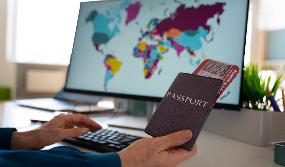 Uma pessoa segura um passaporte e um cartão de embarque em frente a uma tela de computador exibindo um mapa-múndi colorido. A cena sugere planejamento de viagem, possivelmente para imigração laboral. Um teclado repousa sobre a mesa ao lado de um vaso com pequenas plantas verdes, aproveitando a luz natural do ambiente. - Efacont