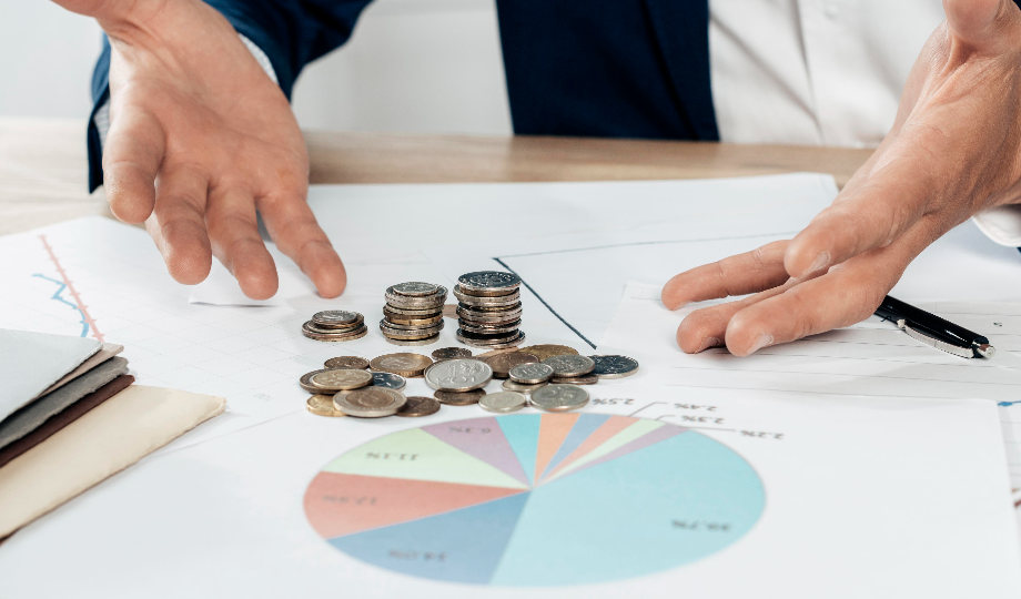 Uma pessoa em traje profissional gesticula com as mãos abertas sobre pilhas de moedas variadas em uma mesa coberta com documentos financeiros, incluindo um gráfico de pizza colorido e um gráfico de linhas, sugerindo planejamento financeiro para os concursos do PT2030. - Efacont