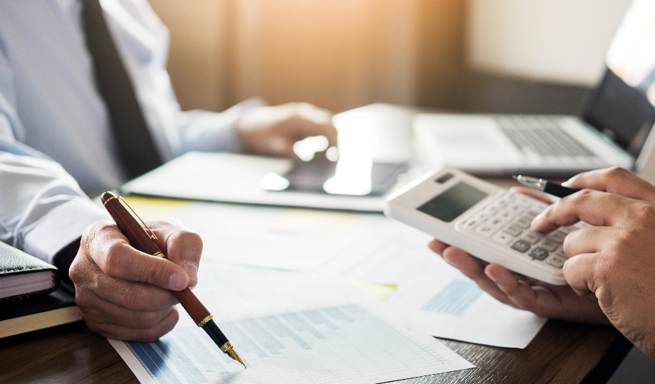 Duas pessoas estão sentadas a uma mesa com documentos, gráficos, um laptop e um caderno. Uma aponta para um gráfico com uma caneta enquanto a outra usa uma calculadora, indicando uma consultoria fiscal ou discussão financeira em andamento. - Efacont