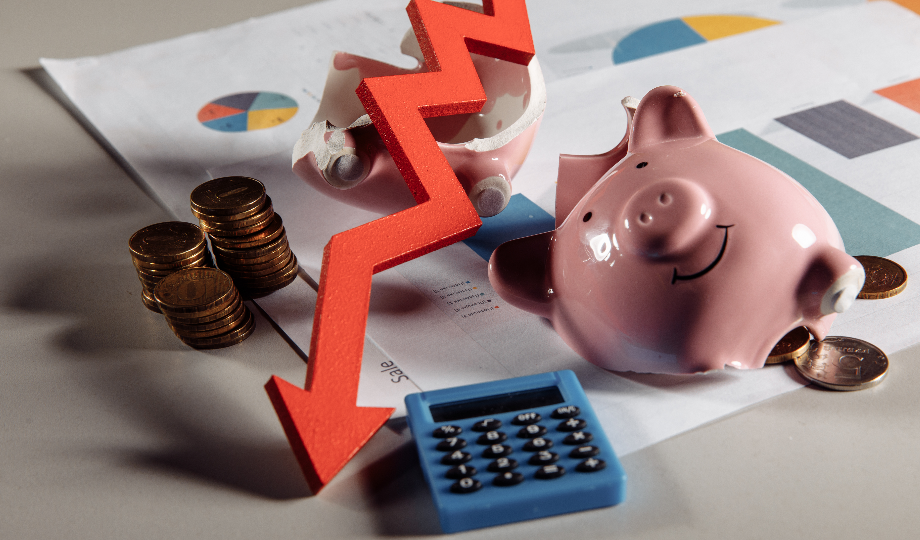 Um cofrinho rosa quebrado repousa sobre gráficos financeiros, ao lado de moedas empilhadas e uma calculadora azul. Uma seta vermelha em zigue-zague apontando para baixo destaca o impacto da perda da estabilidade financeira ou do declínio econômico. - Efacont