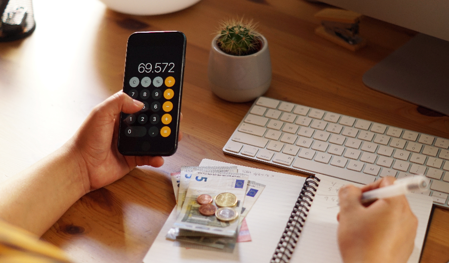 Uma pessoa explora o significado do MTIC enquanto segura um smartphone com um aplicativo de calculadora exibindo 69,572. Sobre a mesa de madeira, há notas de euro, moedas, um bloco de notas, um teclado branco e um pequeno cacto, enquanto escrevem no bloco com uma caneta. - Efacont