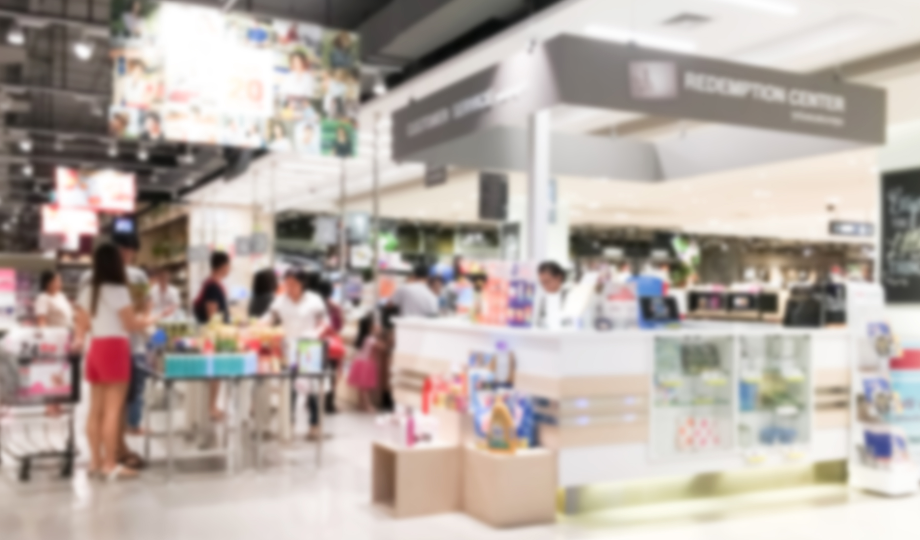 O interior de um supermercado bem iluminado mostra pessoas fazendo compras e formando filas perto de um balcão central de trocas. O cenário movimentado, que lembra grandes eventos empresariais, apresenta prateleiras abastecidas e displays coloridos. A imagem geral está propositalmente desfocada. - Efacont