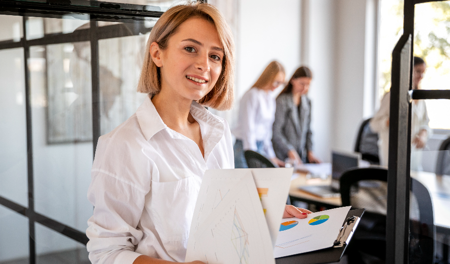 Uma mulher sorridente, de cabelos loiros curtos, segura uma prancheta com gráficos sobre o que é um modelo de negócio em um escritório bem iluminado. Ela veste uma camisa branca, enquanto três colegas colaboram em uma mesa perto de grandes janelas. O ambiente parece profissional e amigável. - Efacont