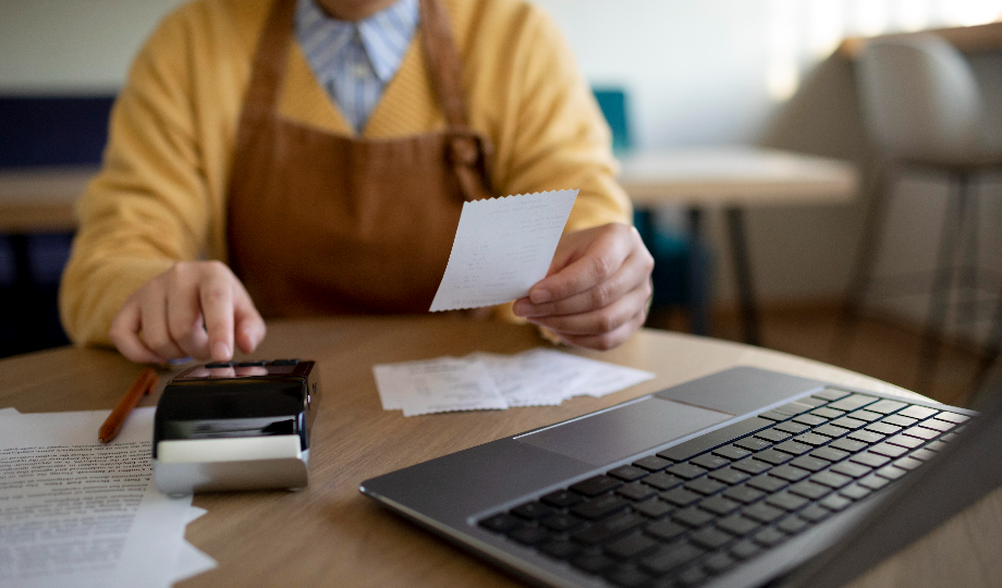 Uma pessoa com um suéter amarelo e avental está sentada à mesa usando uma calculadora, segurando um recibo — talvez revisando o que é nota de crédito — cercada por outros recibos, documentos e um laptop aberto enquanto se concentra em trabalho financeiro ou comercial. - Efacont