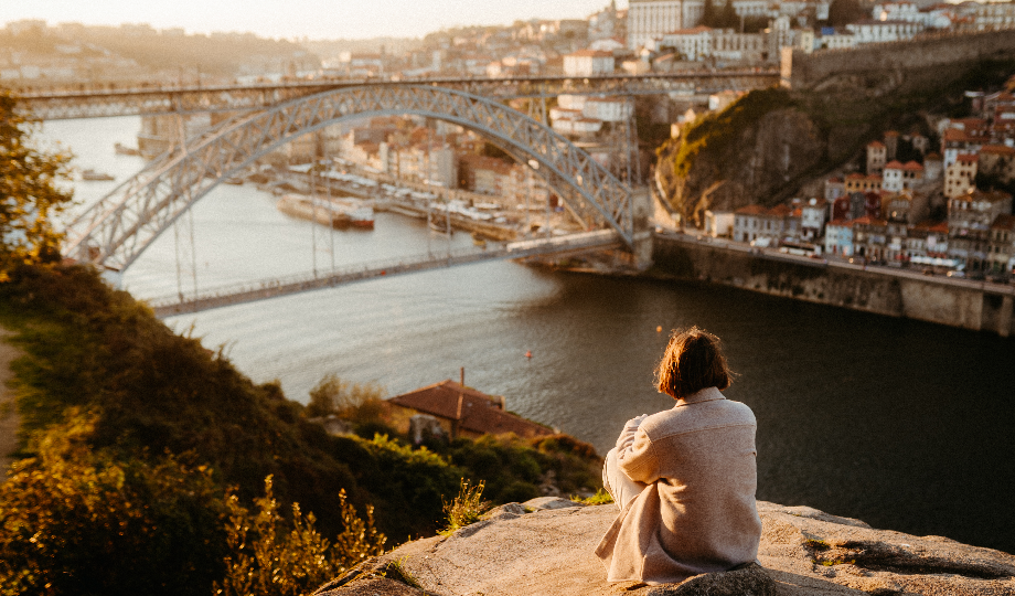 Uma pessoa de cabelo curto e casaco leve senta-se numa saliência rochosa, com vista para o rio e a Ponte Dom Luís I, no Porto. Com o crescimento do turismo em Portugal, o pôr do sol banha os edifícios e a água da cidade com uma luz dourada. - Efacont
